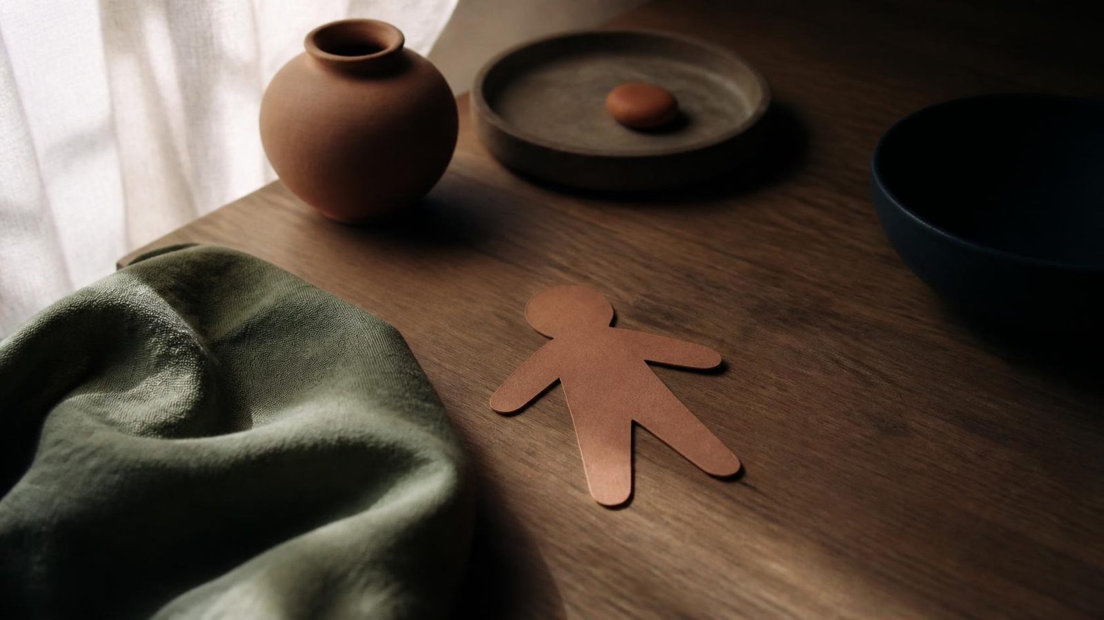 A small flat clay paper figure on an oak desk beside a sage cloth and a clay pebble, in window light.