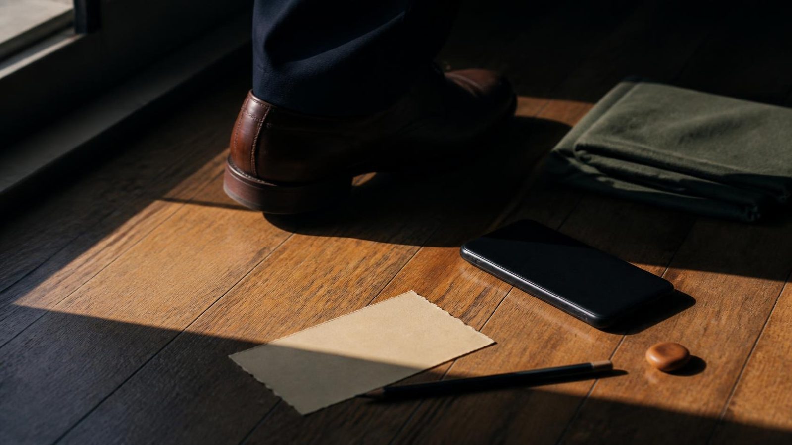 A leather shoe mid-step on oak floorboards, with a face-down phone, blank paper, and a small clay pebble.